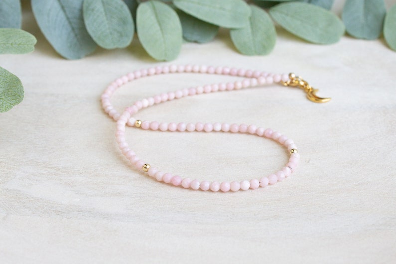 May include: A delicate pink beaded necklace with gold accents. The necklace features small, round pink beads and gold-colored beads. A gold-colored crescent moon charm is attached to the clasp. The necklace is displayed on a light-colored surface with green leaves in the background.