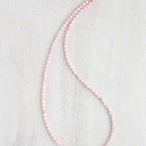 May include: A delicate necklace featuring small, light pink beads strung together. The necklace has gold-colored accents and a gold-colored crescent moon clasp. The beads are evenly spaced, creating a simple yet elegant design.