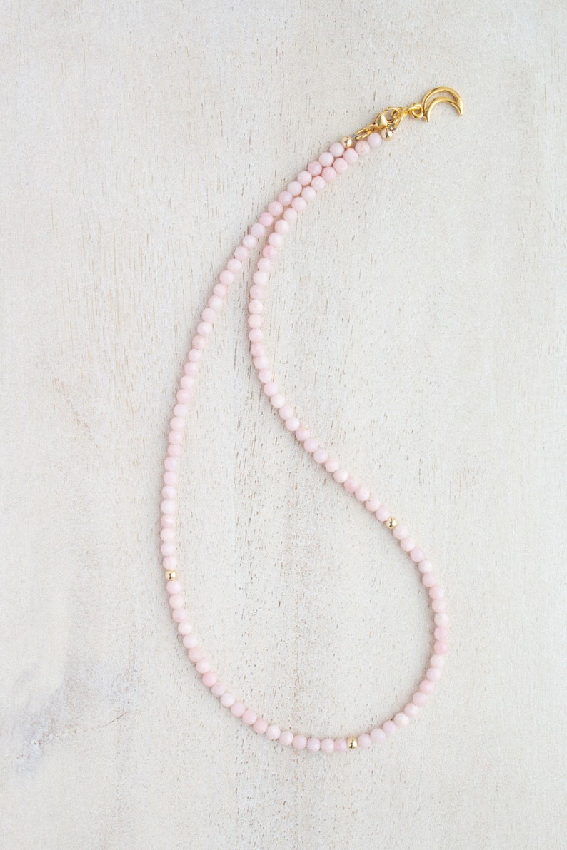 May include: A delicate necklace featuring small, light pink beads strung together. The necklace has gold-colored accents and a gold-colored crescent moon clasp. The beads are evenly spaced, creating a simple yet elegant design.