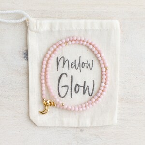 May include: A delicate pink beaded necklace with gold accents and a crescent moon clasp rests on a cream-colored drawstring bag. The bag features the words "Mellow Glow" in a handwritten font. The necklace is a jewelry item.
