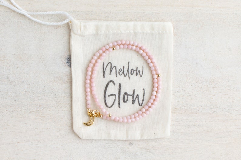 May include: A delicate pink beaded necklace with gold accents and a crescent moon clasp rests on a cream-colored drawstring bag. The bag features the words "Mellow Glow" in a handwritten font. The necklace is a jewelry item.