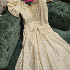 May include: Ivory-colored vintage wedding dress with short, puffed sleeves and a fitted bodice. The dress features decorative floral accents at the waist and a button-down front. The skirt is long and flowing, with vertical beaded lines.