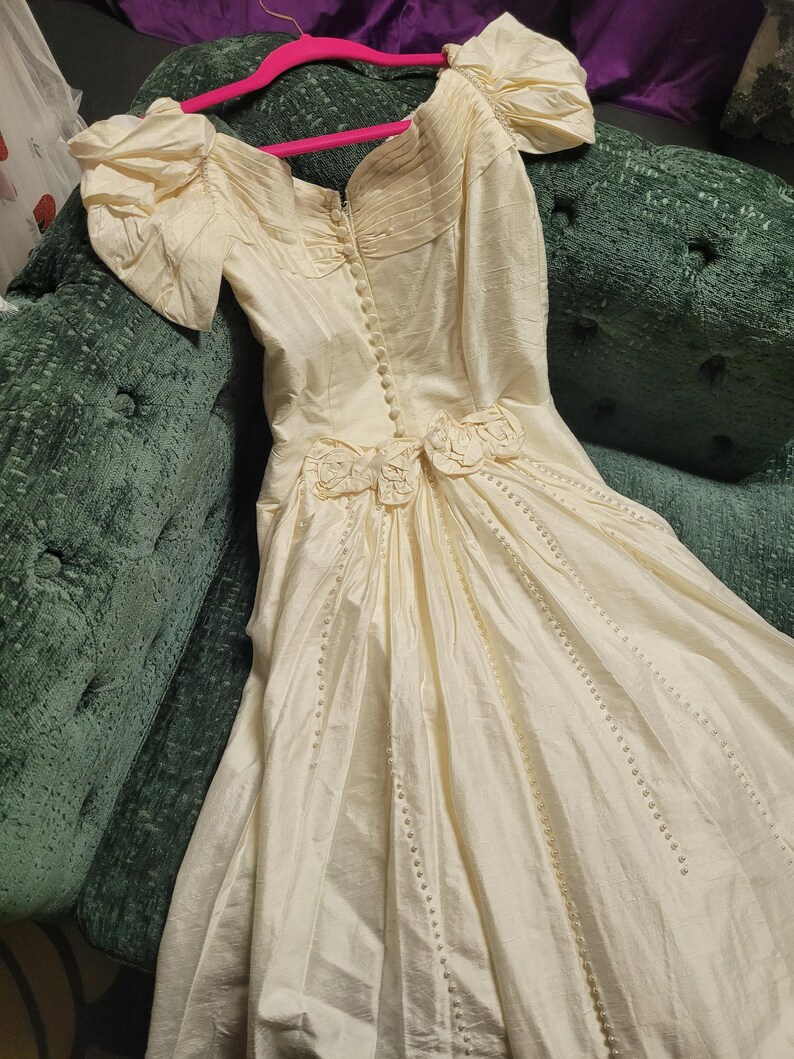 May include: Ivory-colored vintage wedding dress with short, puffed sleeves and a fitted bodice. The dress features decorative floral accents at the waist and a button-down front. The skirt is long and flowing, with vertical beaded lines.