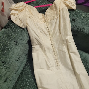 May include: Ivory-colored vintage dress with a sweetheart neckline and puff sleeves. The dress features a row of buttons down the front and a fitted bodice. The fabric has a subtle sheen, suggesting a silk or satin material. The dress is hanging on a pink hanger.