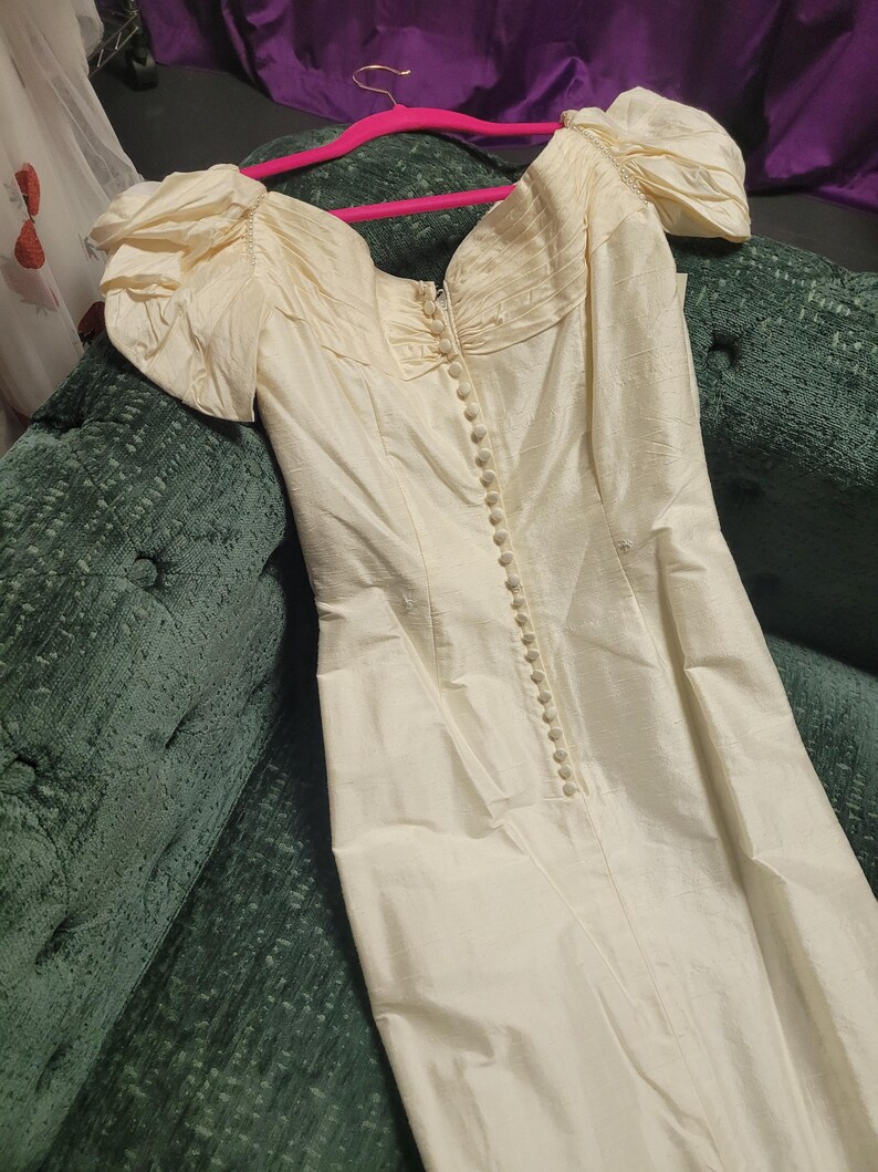 May include: Ivory-colored vintage dress with a sweetheart neckline and puff sleeves. The dress features a row of buttons down the front and a fitted bodice. The fabric has a subtle sheen, suggesting a silk or satin material. The dress is hanging on a pink hanger.