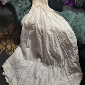 May include: An ivory-colored wedding dress with a long train, displayed on a green velvet sofa. The dress features a V-neckline, short puffed sleeves, and decorative floral accents at the waist. The fabric appears to be a soft, flowing material.