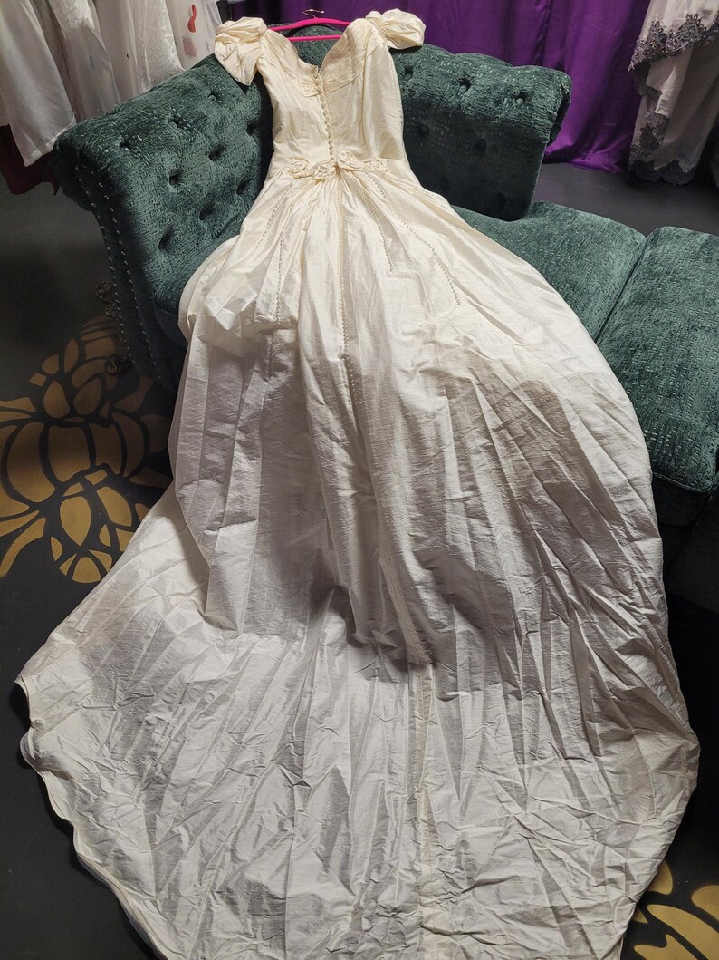 May include: An ivory-colored wedding dress with a long train, displayed on a green velvet sofa. The dress features a V-neckline, short puffed sleeves, and decorative floral accents at the waist. The fabric appears to be a soft, flowing material.