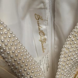 May include: Close-up of a cream-colored wedding dress with a pearl-beaded neckline. The dress features a zipper and a tag with the text "Bridal Originals". The fabric appears to be silk and polyester.