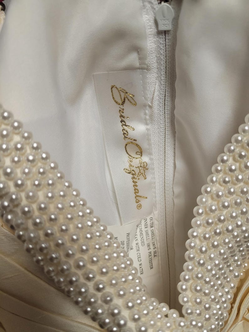 May include: Close-up of a cream-colored wedding dress with a pearl-beaded neckline. The dress features a zipper and a tag with the text "Bridal Originals". The fabric appears to be silk and polyester.