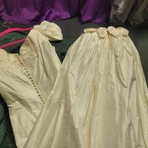 May include: Ivory-colored vintage wedding dress with a bodice featuring a row of buttons and short, puffed sleeves. The skirt is long and flowing, with a decorative floral detail at the waist. The dress is displayed on a green surface.