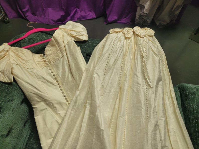 May include: Ivory-colored vintage wedding dress with a bodice featuring a row of buttons and short, puffed sleeves. The skirt is long and flowing, with a decorative floral detail at the waist. The dress is displayed on a green surface.