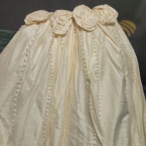 May include: Ivory-colored fabric with a satin finish, adorned with rows of small pearls and floral embellishments. The top edge features several fabric roses. The fabric is draped, suggesting a formal garment.