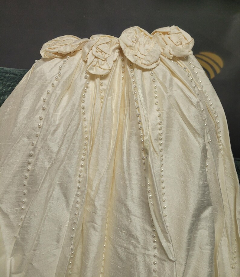 May include: Ivory-colored fabric with a satin finish, adorned with rows of small pearls and floral embellishments. The top edge features several fabric roses. The fabric is draped, suggesting a formal garment.