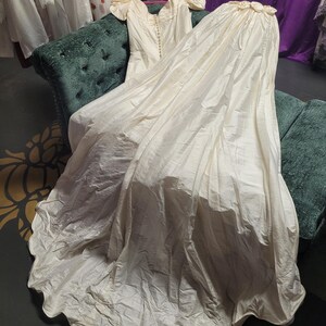 May include: An ivory-colored vintage wedding dress with a long train, resting on a green velvet settee. The dress features a button-down bodice, short sleeves, and a flowing skirt. The train is gathered at the top with floral accents.