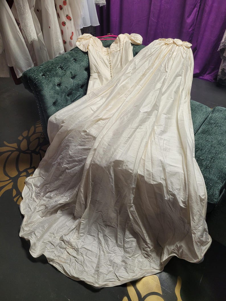 May include: An ivory-colored vintage wedding dress with a long train, resting on a green velvet settee. The dress features a button-down bodice, short sleeves, and a flowing skirt. The train is gathered at the top with floral accents.
