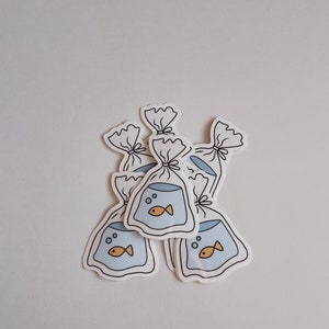 May include: Five white stickers with a cartoon drawing of a goldfish in a plastic bag. The goldfish is orange and has a black outline. The plastic bag is blue and has a white outline. The stickers are arranged in a pile.