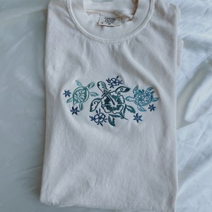 Może przedstawiać: Biały t-shirt z haftowanym wzorem przedstawiającym trzy ż&oacute;łwie morskie i kwiaty. Na metce widnieje napis "Comfort Colors".