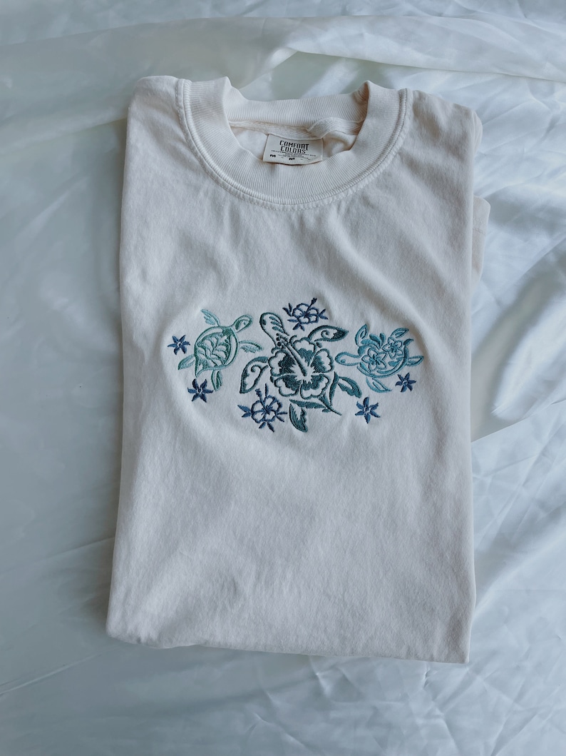 Może przedstawiać: Biały t-shirt z haftowanym wzorem przedstawiającym trzy ż&oacute;łwie morskie i kwiaty. Na metce widnieje napis "Comfort Colors".