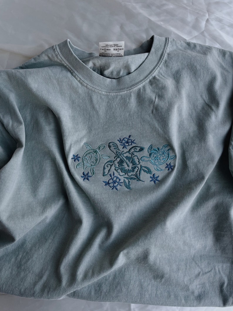 Może przedstawiać: Jasno błękitny t-shirt z haftowanym wzorem trzech morskich ż&oacute;łwi i kwiat&oacute;w. Koszulka ma okrągły dekolt i kr&oacute;tkie rękawy. Etykieta na koszulce głosi "100% Bawełna Wyprodukowano w Hondurasie" i "TADEO TADEO".