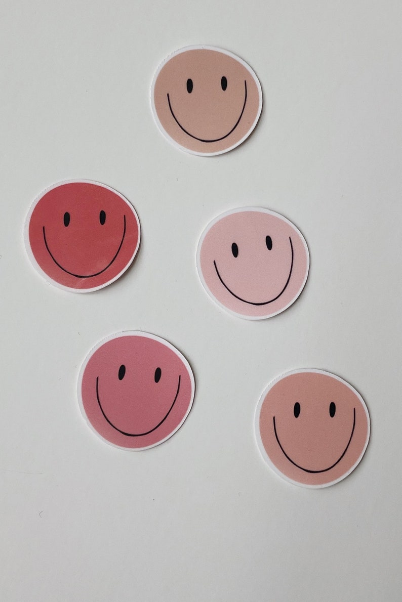 May include: Five smiley face stickers in various shades of pink and peach. The stickers are round and have a black outline.