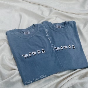 Può includere: Due magliette blu con una fila di pecore bianche ricamate sul davanti. Le magliette hanno il testo "Molly Louise Shop" scritto in corsivo bianco in basso.