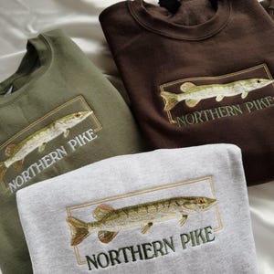 May include: Three sweatshirts in different colors, green, brown, and gray, with an embroidered image of a northern pike fish and the text "NORTHERN PIKE" on each.