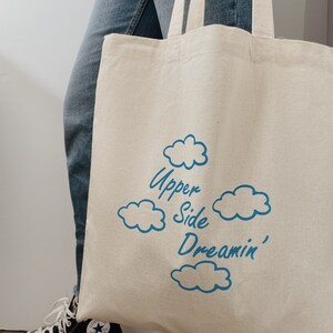 May include: A white canvas tote bag with blue text that reads "Upper Side Dreamin'" and blue cloud illustrations.