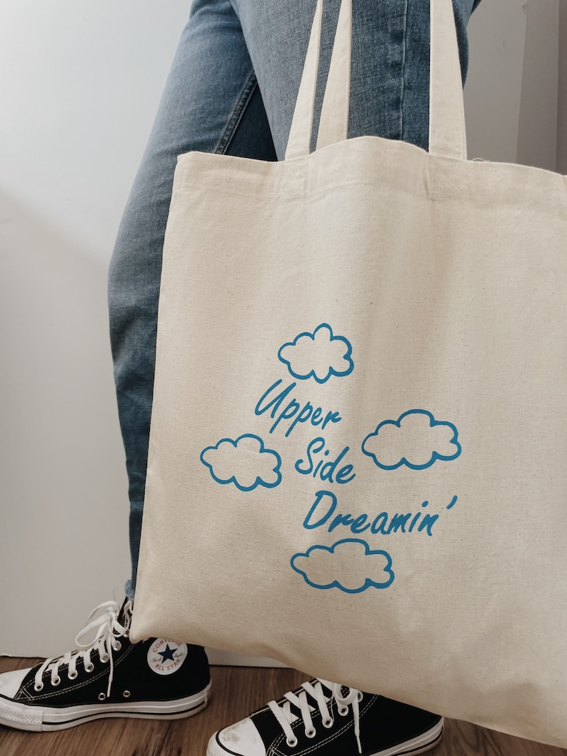 May include: A white canvas tote bag with blue text that reads "Upper Side Dreamin'" and blue cloud illustrations.