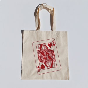 May include: A cream-colored tote bag with a red Queen of Hearts playing card design. The card features a detailed illustration of a queen and heart symbols. The bag has two handles for carrying.