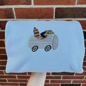 May include: A light blue sweatshirt featuring an embroidered raccoon driving a log-shaped vehicle with wheels. The raccoon has brown and tan fur, green eyes, and is wearing a small hat. The vehicle is gray with black wheels.