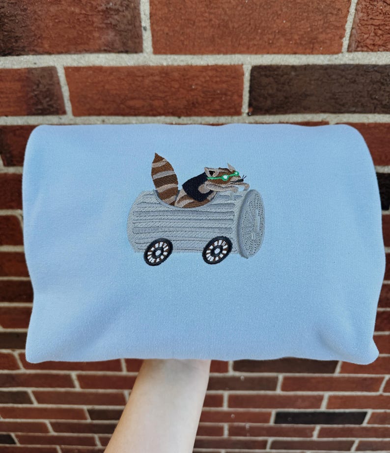 May include: A light blue sweatshirt featuring an embroidered raccoon driving a log-shaped vehicle with wheels. The raccoon has brown and tan fur, green eyes, and is wearing a small hat. The vehicle is gray with black wheels.