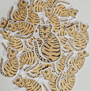 Puede incluir: Una colección de más de 20 pegatinas de tigres, cada una con un dibujo lineal simple de un tigre en posición sentada. Los tigres están todos mirando hacia adelante y tienen una expresión similar. Las pegatinas son todas blancas con contornos negros y están impresas sobre un fondo transparente.