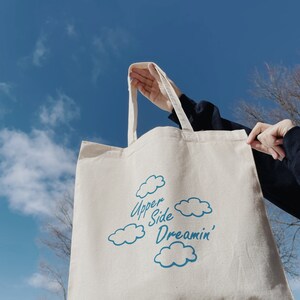 May include: A white canvas tote bag with blue text and cloud designs. The text reads "Upper Side Dreamin'"