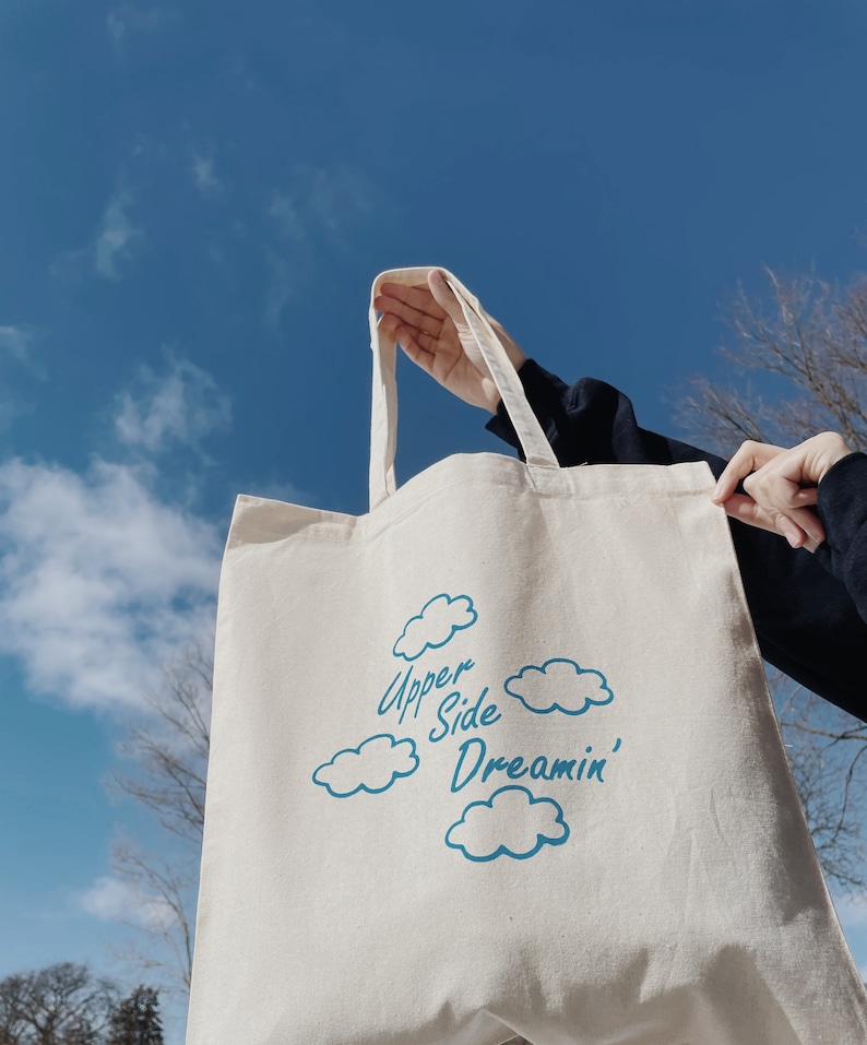 May include: A white canvas tote bag with blue text and cloud designs. The text reads "Upper Side Dreamin'"