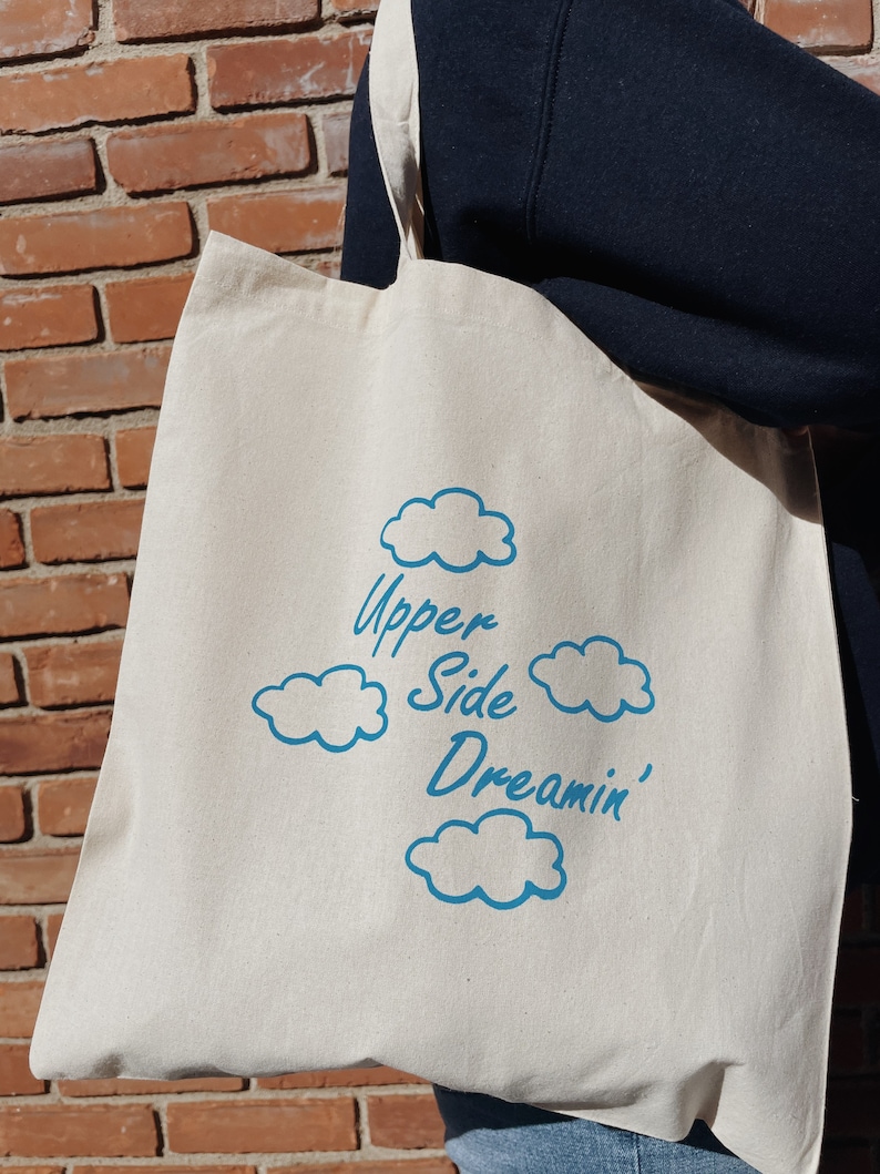 May include: A white canvas tote bag with blue text and cloud designs. The text reads "Upper Side Dreamin'"