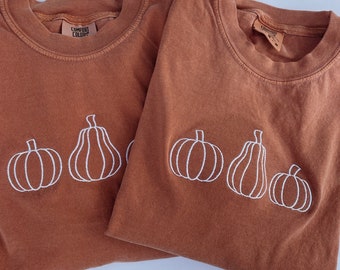 Könnte beinhalten: Zwei rostfarbene T-Shirts mit weißen gestickten Kürbissen. Die Shirts haben den Text "Comfort Colors" auf dem Nackenlabel.