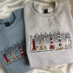 May include: A light blue crewneck sweatshirt and a heather gray crewneck sweatshirt, each featuring an embroidered design of five distinct lighthouses with the text "CLASSIC LIGHTHOUSES" stitched in dark blue thread above the lighthouse illustrations.