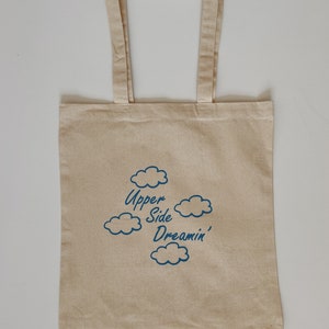 May include: A beige canvas tote bag with blue text and cloud illustrations. The text reads "Upper Side Dreamin'"