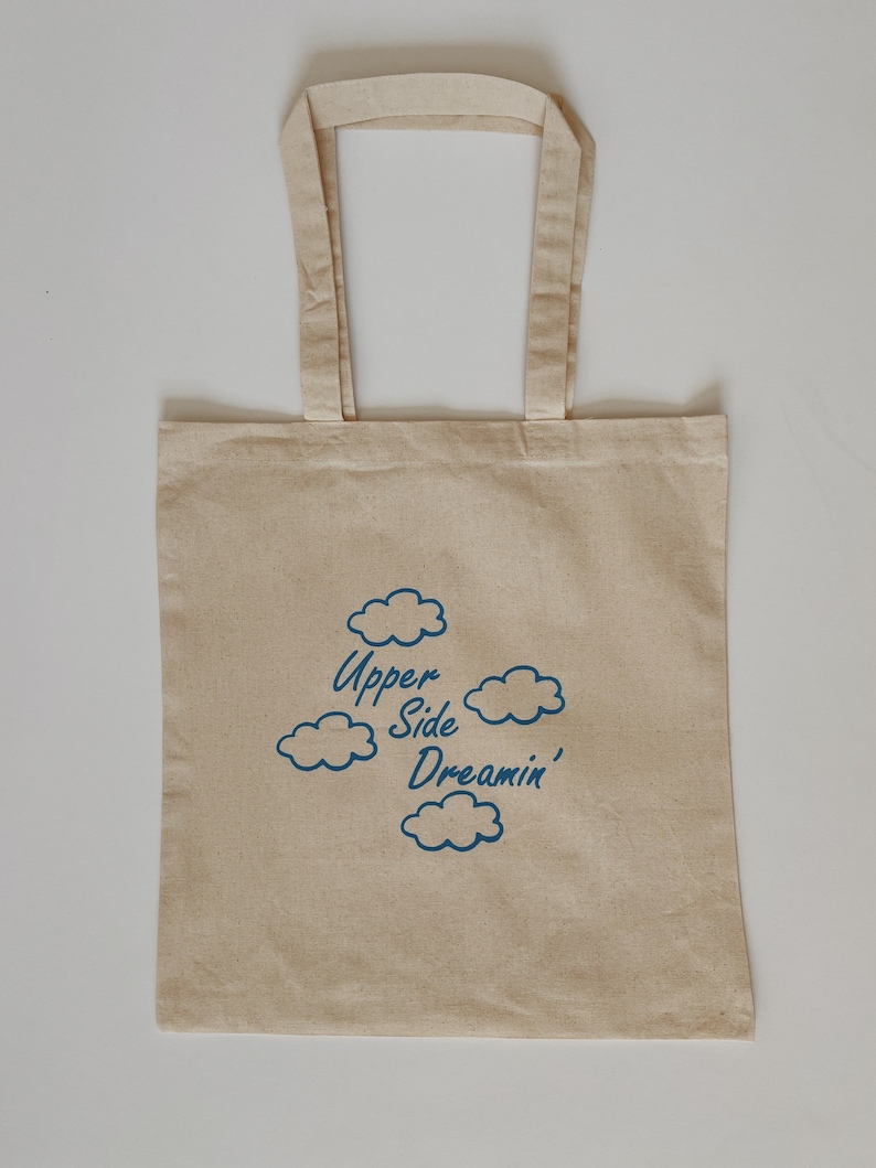 May include: A beige canvas tote bag with blue text and cloud illustrations. The text reads "Upper Side Dreamin'"