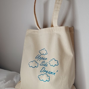 May include: A beige canvas tote bag with blue cloud graphics and the text "Upper Side Dreamin'" printed on the front.