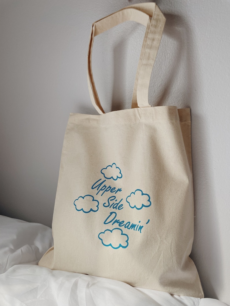 May include: A beige canvas tote bag with blue cloud graphics and the text "Upper Side Dreamin'" printed on the front.