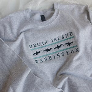 May include: A light grey sweatshirt featuring the text "ORCAS ISLAND WASHINGTON" in a rectangular design. The design includes black orca whale silhouettes and teal stripes. The sweatshirt is made of a soft material.