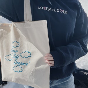 May include: A person wearing a black sweatshirt with the text "LOSER=LOVER" in pink and white lettering. They are holding a white tote bag with the text "Upper Side Dreamin'" in blue lettering and a cloud design.