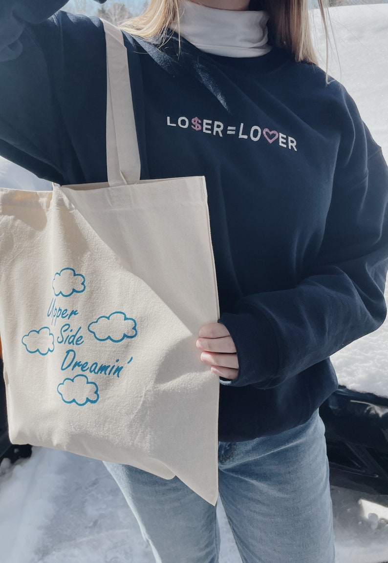 May include: A person wearing a black sweatshirt with the text "LOSER=LOVER" in pink and white lettering. They are holding a white tote bag with the text "Upper Side Dreamin'" in blue lettering and a cloud design.