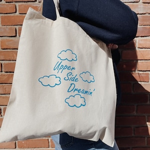 May include: A white canvas tote bag with blue text that reads "Upper Side Dreamin'" and blue cloud illustrations.