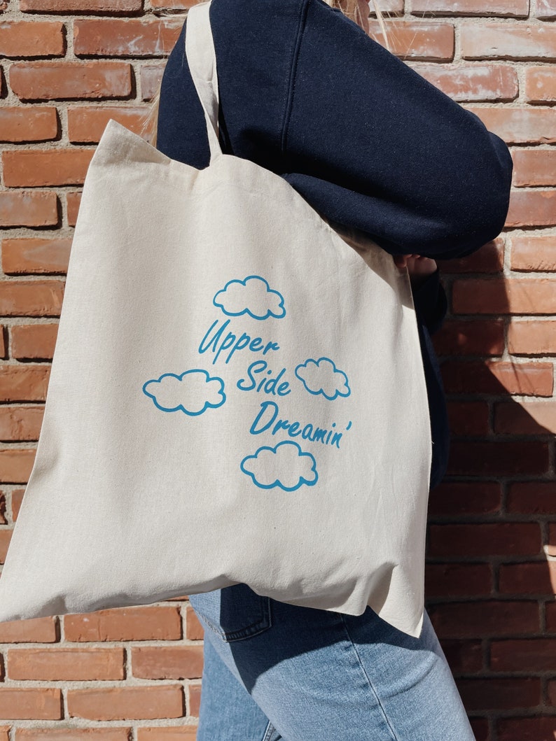 May include: A white canvas tote bag with blue text that reads "Upper Side Dreamin'" and blue cloud illustrations.
