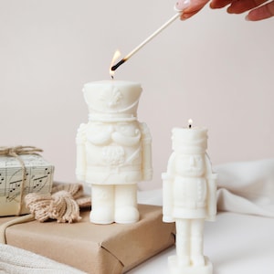 May include: Two ivory-coloured, soldier-shaped candles, one being lit with a match. The candles are detailed with uniform features. A gift wrapped in brown paper and a white sweater are in the background. The scene is set on a white surface.