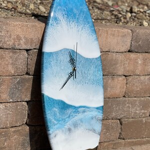 May include: A blue and white surfboard clock with a black clock face and hands. The surfboard is painted to look like ocean waves.