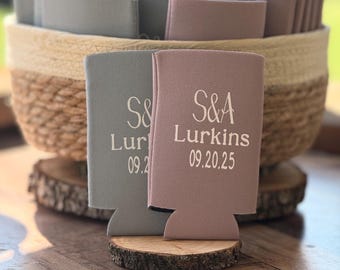 May include: Two personalized can coolers, one gray and one mauve, with white text reading "S&A Lurkins 09.20.25." A basket filled with similar coolers sits in the background. The coolers are displayed on wooden slices, suggesting a rustic theme.