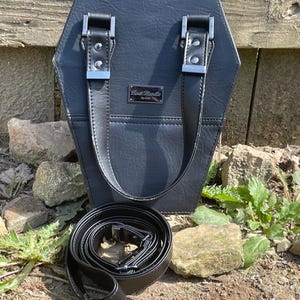 May include: A black coffin-shaped handbag with a textured surface, two fixed top handles with silver-tone metal hardware, and a detachable shoulder strap coiled at the base. A rectangular silver metal plaque features the text "Lost Needle by Azaz".